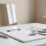 A minimalist medical consultation desk featuring a sleek stethoscope, a clipboard with blank charts, and a pen, bathed in soft natural light with a simple mirror in the background reflecting an empty beige wall.